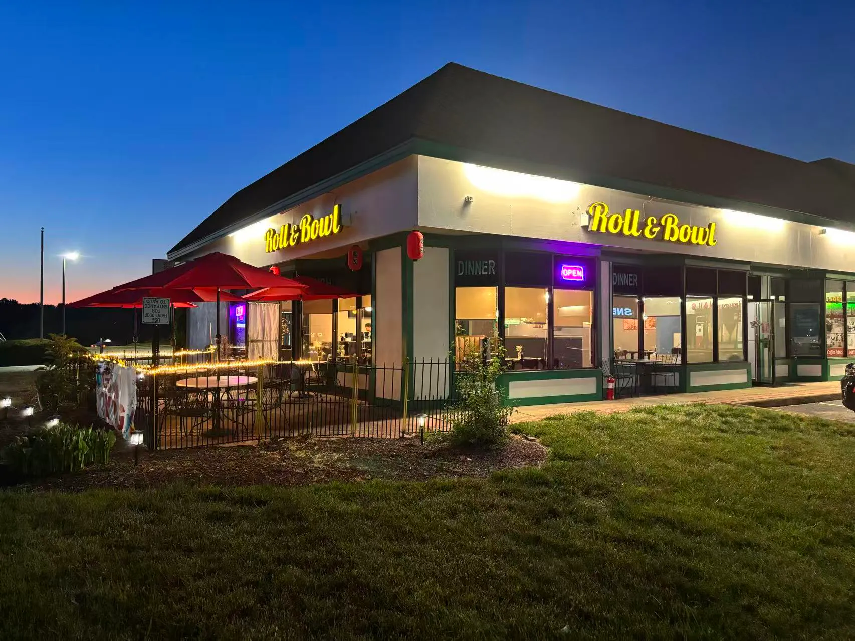Exterior of Roll & Bowl restaurant at dusk with a lit outdoor patio and red umbrellas.